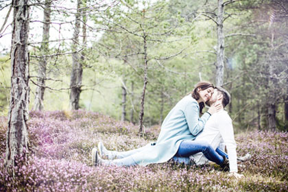 Verliebtes Paar im Frühling auf einer Waldlichtung sitzend - Er flüstert ihr etwas ins Ohr - sie lächelt und lehnt ihr Gesicht an seinen Hals Sexualberatung für Mölln